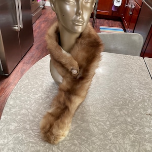 Puede incluir: Una estola larga de piel marrón con vientre blanco. La estola está drapeada alrededor del cuello de un maniquí y tiene un broche decorativo con una piedra transparente en el centro.