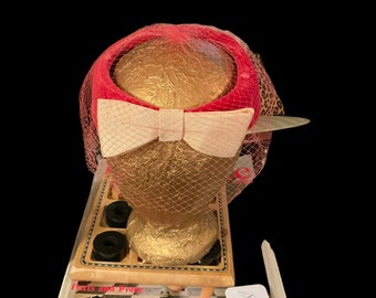 May include: A pink and white pillbox hat with a net veil. The hat is sitting on a vintage book titled "Paris and Provence 175 Fashion Pages in Full Color Photography".