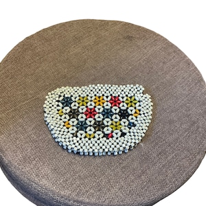 May include: A round brown cushion with a colorful beaded seat cushion. The beaded cushion is made of white beads with red, yellow, blue, and black accents.
