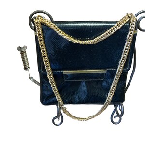 May include: A black, textured handbag with a gold chain strap. The bag has a rectangular shape with a flap closure and a gold bar detail. The bag is displayed on a decorative metal stand with curled accents.