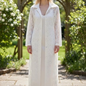 May include: A white lace duster coat with long sleeves and a collared neckline. The coat is worn over a white dress. The image is taken outdoors in a garden setting with a stone pathway and greenery in the background.