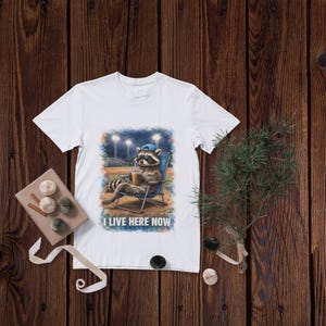 Può includere: T-shirt bianca con un procione in uno stadio di baseball, con un berretto blu e seduto su una sedia a sdraio. Il procione tiene in mano una bevanda. Il testo "I LIVE HERE NOW" è stampato sotto.
