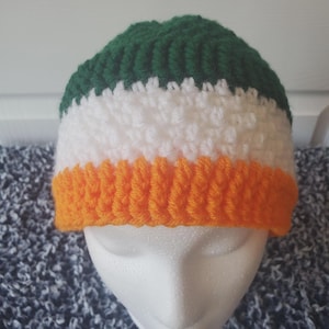 Könnte beinhalten: Eine gestrickte Mütze mit dreifarbigem Design. Die Mütze hat horizontale Streifen in Grün, Weiß und Orange. Die Mütze wird auf einem weißen Schaufensterkopf präsentiert.