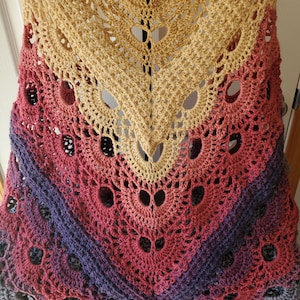 May include: A handmade crochet shawl in a V-shape, featuring a gradient of colors. The top is a light tan, transitioning to pink, purple, and gray at the bottom. The shawl has an open, lacy design with heart-shaped cutouts.
