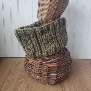 May include: An olive green crocheted cowl featuring a thick, textured cable knit pattern. The cowl is displayed wrapped around a brown woven wicker basket, which is positioned on top of a larger, rounded brown woven wicker base structure.
