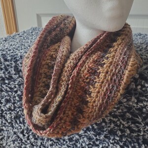 May include: A chunky crochet infinity scarf featuring a variegated yarn blend of warm earth tones including shades of brown, tan, rust, and deep burgundy. The scarf has a thick, textured knit pattern and is draped in a circular loop shape.