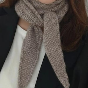 Könnte beinhalten: Ein hellbrauner Strickschal mit einer strukturierten kraus rechts gestrickten Oberfläche. Der Schal ist mit einem einfachen Knoten um den Hals gebunden und hat zwei spitz zulaufende Enden, die nach unten hängen. Das Gestrick wirkt weich und flauschig.
