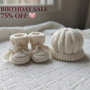 Può includere: Un paio di scarpine per neonati in maglia color crema con lacci e nappe abbinate, insieme a un berretto per neonati in maglia color crema coordinato. Il testo BIRTHDAY SALE 75% OFF con un cuore rosa è visibile in alto nell'immagine.