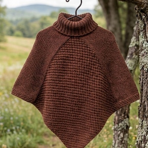 Può includere: Un poncho in maglia marrone scuro con collo alto a coste e un motivo a nido d'ape testurizzato sul pannello frontale. Il capo è appeso a una gruccia in metallo nero e presenta un orlo a punta e maniche in maglia liscia.