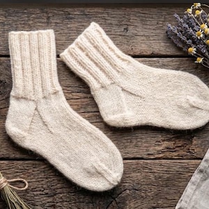 May include: A pair of cream-coloured knit socks featuring a ribbed cuff design and a smooth, plain-knit foot section. The socks are crafted with a visible textured yarn, showcasing a classic construction with a reinforced heel and toe area.
