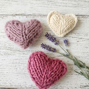 Könnte beinhalten: Drei gestrickte herzförmige Ornamente in Rosa- und Cremetönen, angeordnet mit Lavendelzweigen auf einer weißen Holzoberfläche. Die Herzen haben ein strukturiertes, handgemachtes Aussehen mit sichtbaren Garnstichen. Der Lavendel fügt einen Hauch von natürlicher Farbe hinzu.