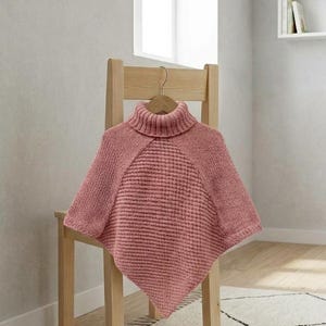 Könnte beinhalten: Ein altrosa gestrickter Poncho mit hohem Rollkragen, einem strukturierten Waffelstrick-Mittelteil und glatten Seitenpartien in Glattstrickoptik. Das Kleidungsstück hängt an einem Holzkleiderbügel und zeigt einen spitz zulaufenden Saum.
