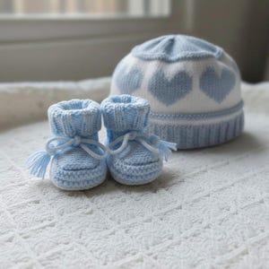 Op de afbeelding: Een paar lichtblauwe gebreide babyslofjes met witte trekkoorden en kwastjes, vergezeld van een bijpassend lichtblauw en wit gebreid babymutsje met een herhalend hartjespatroon rond de basis en een geribbelde boord.