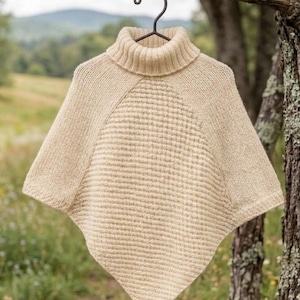 Könnte beinhalten: Ein cremefarbener Strickponcho mit hohem Rollkragen und einem strukturierten Waffelstrickmuster auf der Vorderseite. Das Kleidungsstück ist mit glatten Strickärmeln und Seitenteilen gefertigt und hängt an einem schwarzen Metallbügel mit Bindfaden.