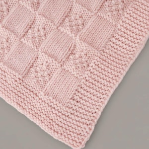 Puede incluir: Primer plano de una manta de punto rosa suave. La manta presenta un patrón texturizado de cuadrados y rectángulos, con un diseño de puntada detallado dentro de algunos de los cuadrados. Los bordes están acabados con un borde acanalado, creando una apariencia acogedora.