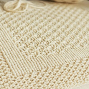 Può includere: Coperta lavorata a maglia color crema con un motivo strutturato. La coperta presenta un bordo a coste e un design a punto dettagliato, che mostra un'estetica fatta a mano. Un gomitolo di lana è visibile.