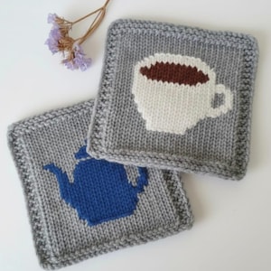 May include: Two square knitted coasters in grey yarn. One coaster features a white coffee cup with dark brown liquid inside. The other coaster displays a solid blue teapot silhouette. Both items have a garter stitch border and a smooth stockinette centre.