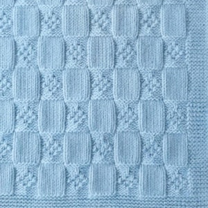 May include: A light blue knitted blanket featuring a repeating pattern of smooth stockinette stitch squares alternating with textured seed stitch squares. The blanket is finished with a wide garter stitch border along the bottom edge.