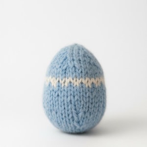 May include: A pale blue knitted egg with a white horizontal stripe. The egg-shaped item is made of soft yarn, possibly for decoration or as a toy. The background is plain white.