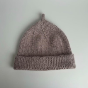 Może przedstawiać: Dzianinowa czapka beanie w stonowanym kolorze mauve. Czapka ma składany brzeg i spiczasty wierzch z małym, pionowym przedłużeniem. Widoczna jest faktura dzianiny, co sugeruje ręczną lub rzemieślniczą jakość.