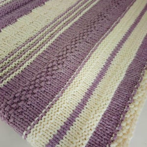 May include: A knitted blanket with alternating stripes of lavender and cream. The blanket features textured patterns and a soft, cozy appearance. The design includes diagonal stripes and textured sections.