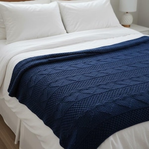 May include: A dark navy blue knitted throw blanket featuring a textured cable knit pattern with diamond shapes and seed stitch details. The blanket is draped over a white bedspread, showcasing the intricate yarn work and deep blue color.