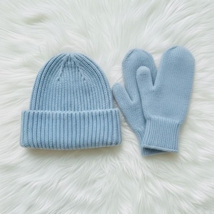 Puede incluir: Un conjunto de gorro y mitones de punto azul claro. El gorro tiene un diseño acanalado con un borde doblado. Los mitones también son acanalados y tienen una forma clásica. El conjunto se muestra sobre una superficie blanca y esponjosa.