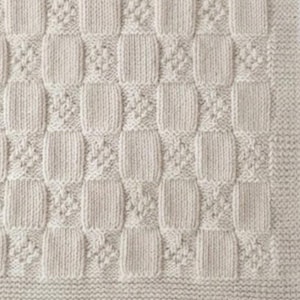 Può includere: Una coperta lavorata a maglia color crema caratterizzata da un motivo ripetuto di quadrati a maglia rasata alternati a quadrati testurizzati a punto nocciolina a forma di diamante, rifinita con un ampio bordo a legaccio sul fondo.