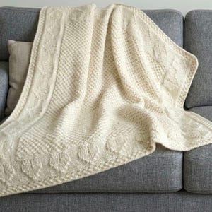 May include: Cream-colored knitted blanket with a heart-shaped border and diamond pattern. The blanket is draped over a gray sofa, showcasing its texture and design. A cozy home decor item.