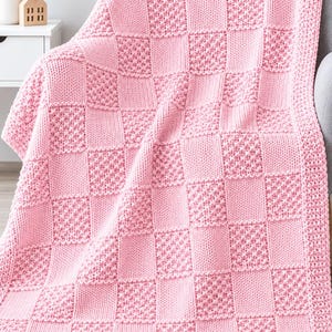 May include: A light pink knitted throw blanket featuring a checkerboard pattern. The design alternates between smooth stockinette stitch squares and textured seed stitch squares, creating a tactile grid across the entire surface of the blanket.