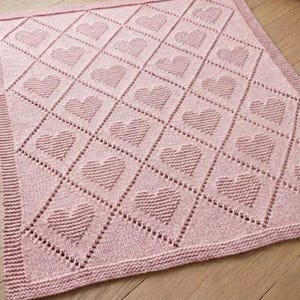 Op de afbeelding: Een lichtroze gebreide babydeken met een herhalend ruitpatroon, waarbij in elke ruit een textuurvorm van een hart is verwerkt. De deken is afgewerkt met een eenvoudige rand in ribbelsteek langs alle vier de zijden van het breiwerk.