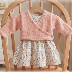 May include: A long-sleeved, light pink knitted wrap cardigan with a single wooden button closure, layered over a white dress featuring a delicate floral pattern with small blue, yellow, and green flowers and a tiered, ruffled skirt hemline.