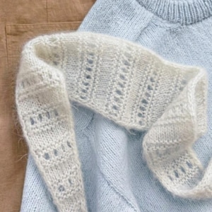 Op de afbeelding: Een lichtblauwe gebreide trui met een geribbelde ronde hals, gecombineerd met een witte pluizige sjaal met een opengewerkt kantpatroon en een beige broek. De kledingstukken zijn over elkaar gelegd en tonen de contrasterende texturen.