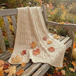 Può includere: Una coperta lavorata a maglia color crema caratterizzata da un motivo a intreccio testurizzato con un bordo a punto riso. La coperta è drappeggiata su una panca in legno ed è decorata con diverse foglie autunnali cadute nei toni dell'arancio e del rosso.