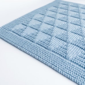 Puede incluir: Una manta tejida de color azul claro con un patrón de diamantes texturizado creado con puntos en relieve. El tejido cuadrado tiene un borde uniforme en punto bobo que enmarca el diseño geométrico central, mostrando un color de hilo constante.