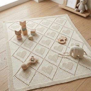 May include: Cream-colored knitted baby blanket with a heart pattern, surrounded by wooden toys, including blocks, a rattle, and a teething ring. A small stuffed elephant is also present. The blanket is approximately 40 inches square.
