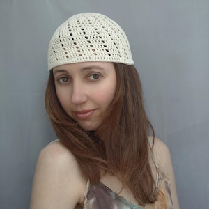 May include: Cream-colored crocheted beanie hat with an open weave design. The hat is a skull cap style, suitable for various occasions. The hat is shown on a person with long brown hair.