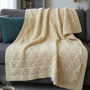 May include: A cream-colored knitted throw blanket featuring a repeating geometric triangle pattern and a textured garter stitch border. The blanket is draped over a dark gray sofa, showcasing the intricate yarn work and soft, neutral tone.