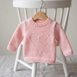 May include: A light pink long-sleeved knit sweater featuring a textured heart pattern on the front. The garment has a ribbed crew neckline, ribbed cuffs, and a ribbed hem, all crafted in a soft, solid-colored yarn with a consistent knit stitch.