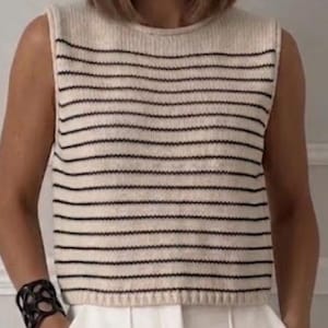 Könnte beinhalten: Eine ärmellose Strickweste mit cremefarbener Basis und dünnen, horizontalen schwarzen Streifen. Das Kleidungsstück hat einen Rundhalsausschnitt und einen gerippten Saum, was für eine strukturierte Passform sorgt. Das Design ist schlicht und betont den Kontrast zwischen dem hellen Stoff und dem dunklen linearen Muster.