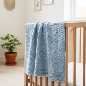 Puede incluir: Una manta de bebé tejida en color azul claro con un patrón texturizado de formas de corazón alternas y pequeños puntos en relieve. La manta está colocada sobre los listones de madera de una cuna, mostrando un color suave y un tejido detallado.