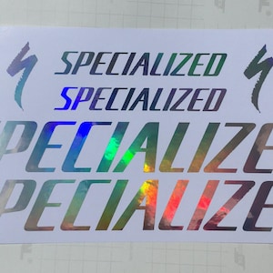 May include: A white decal sheet featuring four Specialized brand logos and text in a holographic finish. Two small lightning bolt icons flank the top text, while two larger, elongated Specialized word marks are positioned below in a reflective rainbow.