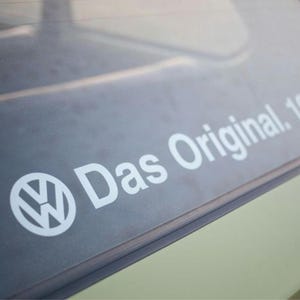 Può includere: Un adesivo in vinile bianco applicato sul finestrino di un veicolo con il logo Volkswagen seguito dal testo Das Original. 1976. L'adesivo è posizionato su una superficie in vetro oscurato con un telaio in metallo verniciato verde chiaro.