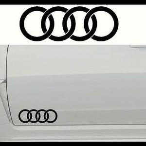 May include: Black interlocking rings logo, a symbol of the Audi brand, displayed on a white car door. The logo is repeated, with one set at the top and another near the bottom of the door.