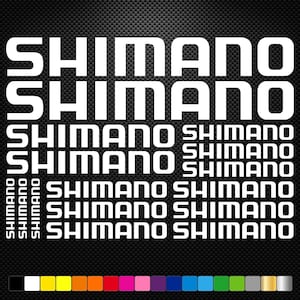 May include: A collection of white Shimano brand decals in various sizes arranged on a black carbon fiber patterned background, with a row of colored square swatches including black, white, yellow, orange, pink, purple, blue, green, and silver.