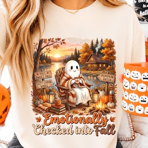 Pode incluir: Uma camisola cor de creme com um gráfico de um fantasma sentado numa cadeira junto a um lago, uma cabana e uma fogueira. O texto diz "FALL SWEET FALL", "COZY CABIN VIBES", "PUMPKIN PATCH" e "Emotionally Checked into Fall" em laranja e castanho.