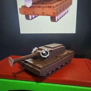 May include: A brown toy tank with cartoon eyes and a long barrel, placed on a red surface. A similar image is displayed on a screen behind it. The tank has a whimsical design, with a playful aesthetic.