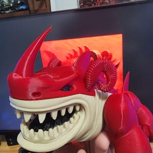 May include: A red and cream-colored 3D-printed figurine of a horned creature with sharp teeth. The figurine has a menacing expression and is held in a person's hand. The creature has a red body, horns, and claws, with a cream-colored face and teeth.