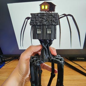 Pode incluir: Uma escultura preta impressa em 3D de uma casa sobre pernas. A casa tem dois andares com janelas e um telhado texturizado. Uma casa menor fica no topo com janelas amarelas brilhantes. A escultura é segurada em uma mão.