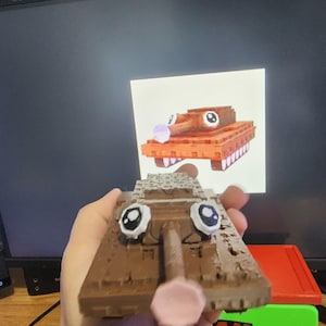 May include: A brown toy tank with cartoon eyes and a pink nose. The tank has a detailed design and is held in front of a computer screen displaying a similar image. The tank is a fun, whimsical item.